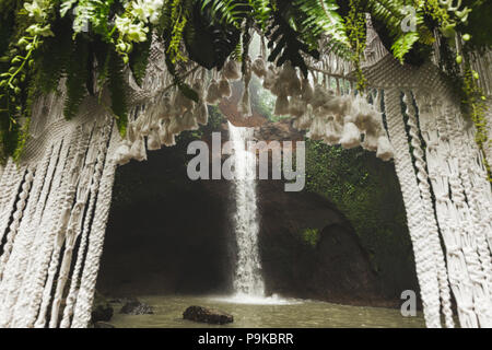 Tropical wedding ceremony with waterfall view. White arch decorated with green jungle leaves monstera and fern Stock Photo