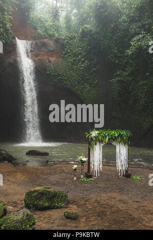 Tropical wedding ceremony with waterfall view. White arch decorated with green jungle leaves monstera and fern Stock Photo