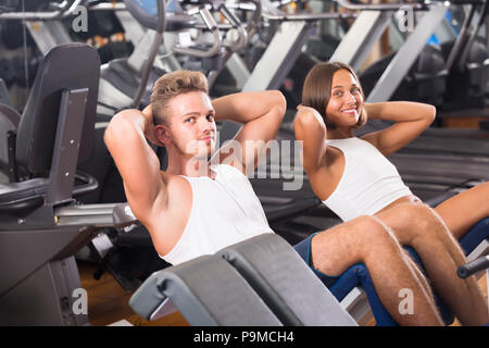 Well trained happy young man and woman training abdominal muscles doing sit ups in gym Stock Photo