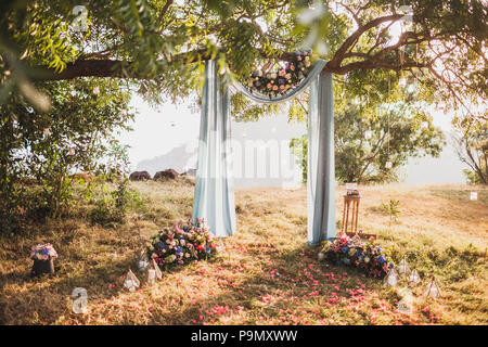 Sunset wedding ceremony, arch decorated with grey cloth hanging on big tree and rose flowers arrangement Stock Photo
