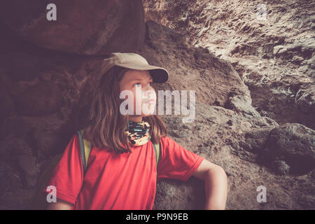 A girl child stares thoughtfully in the middle of a long hike, sheltered under some rocks on a mountain side. She wears a uv scarf, cap, sun cream. Stock Photo