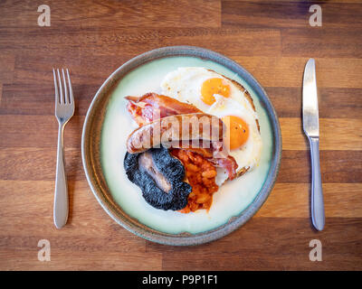 A full English Breakfast with bacon, egg, sausages, beans and mushrooms on a plate on a wooden table with a knife and fork Stock Photo