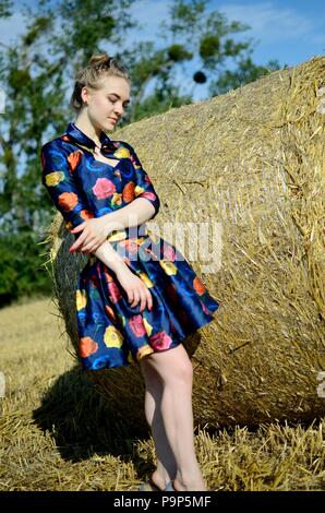 Female model, outdoor photo session, farmland in summer. Girl in colorful dress stands in front of roll of hay. Stock Photo