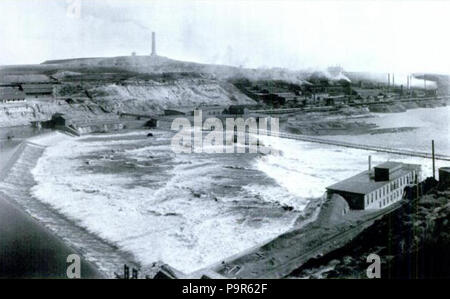 . English: Looking northeast at Black Eagle Dam on the Missouri River in Great Falls, Montana, United States. The original powerhouse built on the south bank of the river is just visible at bottom-center. The second powerhouse, built in 1893-1894 just downstream of the existing south bank powerhouse, is center-left. (Notice the large penstock rising up out of the ground to feed water through the wall of the powerhouse.) The dam itself is in the left of the image. At the dam's northern abutment is the original north bank powerhouse. Downstream of that, just behind the falls and sitting on what  Stock Photo