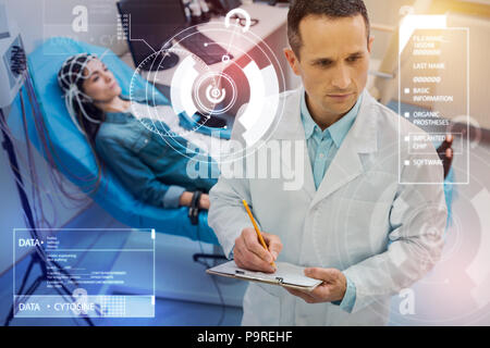 Serious doctor. Attentive qualified doctor making necessary notes while a young woman underdoing electroencephalography behind his back Stock Photo