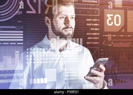 Standing with a smartphone. Calm bearded programmer thoughtfully looking at the transparent screen of his futuristic computer while standing in front  Stock Photo