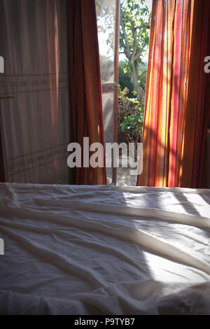 looking over an unmade bed with white sheets out through colourful curtains and an open door onto a garden one sunny morning Stock Photo