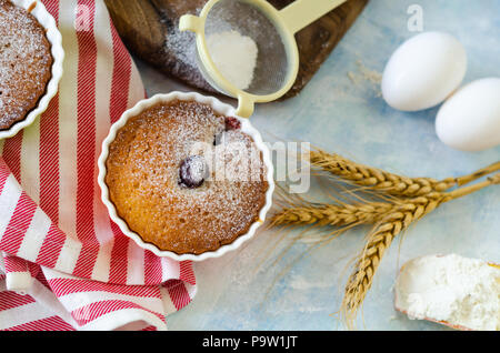 Cakes with cerries on a table in small porcelain cups .There are  eggs, flour, wheat, powdered sugar next to cakes. Stock Photo