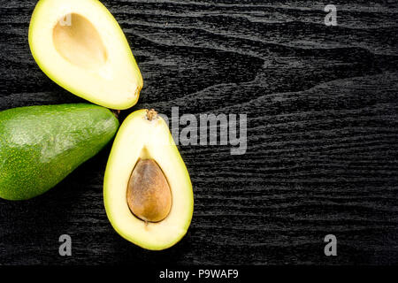 Green smooth avocado table top isolated on black wood background bacon variety one whole two sliced halves with a seed Stock Photo