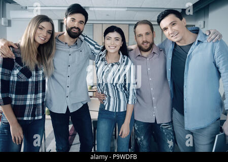 Team spirit. Smiling young group of colleagues bonding to each other and stanting in the office while feeling happy to work together Stock Photo