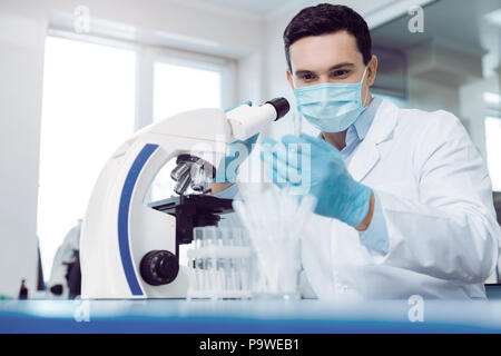 Biological lab. Handsome smart young biologist sitting near the microscope and doing the research while working in the biological lab Stock Photo