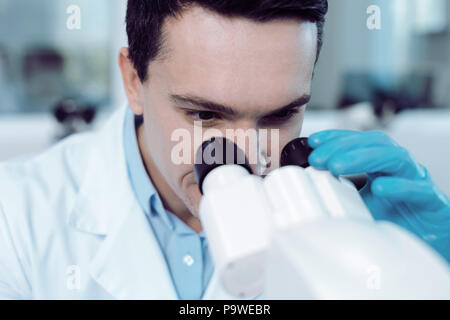 Biological studies. Cheerful positive handsome scientist holding microscope binoculars and looking into them while working on his studies
