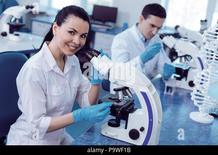 Modern genomics. Delighted positive female geneticist smiling and looking at you while working in the lab with the microscope Stock Photo