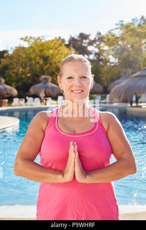 Smiling senior woman folding her hands while doing yoga meditation at the hotel pool Stock Photo