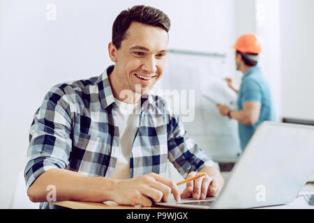 Happy employee. Smiling cheerful positive young worker looking at the screen of a laptop while being in his office Stock Photo