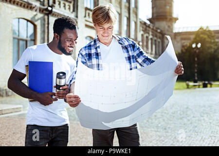 Show it to me. Smiling international student bowing head while looking downwards and standing near his partner Stock Photo