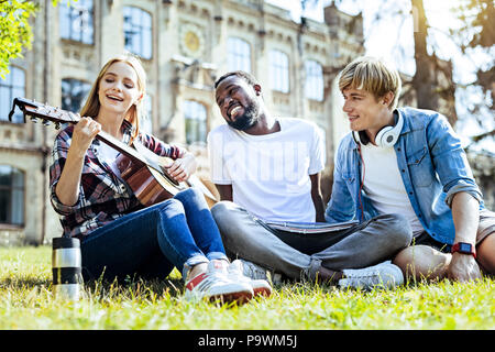 Time to relax. Low angle shot of relaxed young people in casual smiling while listening to their friend playing guitar and singing. Stock Photo