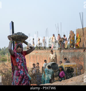 A lifestyle photo of an Indian woman carrying a basket on her head with village workers on a building site in the background Stock Photo