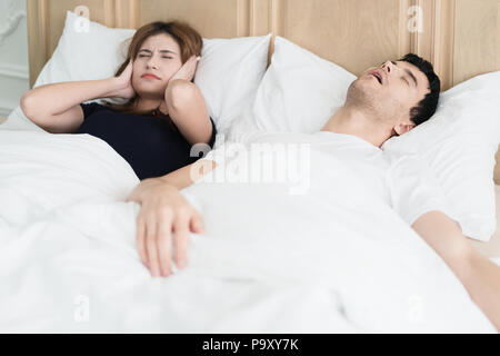 Annoyed Asian wife blocking her ears from noise of husband snoring in bedroom at home. Young couple have problem with man's snoring. Stock Photo