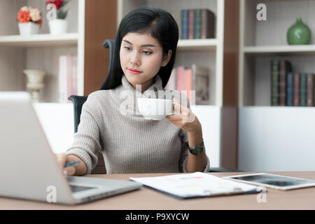 Young Asian businesswoman sitting in business office at desk, drinking coffee and using laptop. On table is laptop. businesswoman browsing internet, c Stock Photo