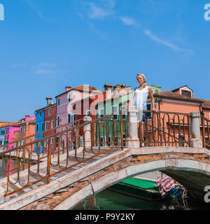 A female traveler stands on a bridge across a canal on the island of Burano with many colorful houses in the background Stock Photo