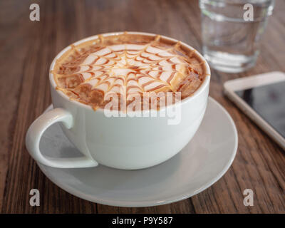 Beautiful Caramel macchiato art on top. Hot coffee in white cup, a glass of water and white mobile phone on wooden background. Stock Photo