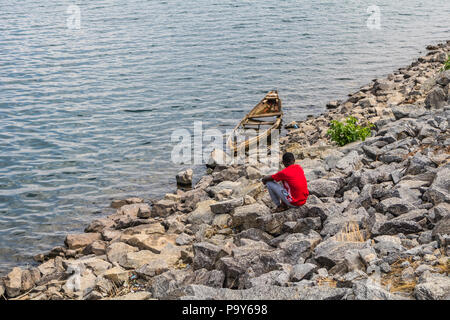 A young man sitting alone and looking at a damaged wooden fishing canoe at the riverbank. Stock Photo