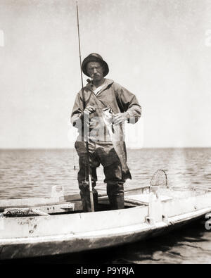 1920s MAN BARNEGAT BAY COMMERCIAL FISHERMAN STANDING IN DUCK BOAT SMOKING PIPE REMOVING FISHHOOK FROM CATCH NEW JERSEY USA - a121 HAR001 HARS SUCCESS REMOVING NJ OCCUPATIONS DUCK BOAT FISHING ROD NEW JERSEY BARNEGAT BAY BLACK AND WHITE CAUCASIAN ETHNICITY HAR001 OLD FASHIONED Stock Photo