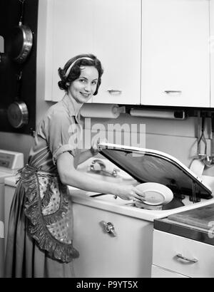 1950s 1960s SMILING HOUSEWIFE LOADING DISHWASHER LOOKING AT CAMERA - bx018415 CAM001 HARS CAM001 HOUSEWIVES SMILES JOYFUL MID-ADULT MID-ADULT WOMAN YOUNG ADULT WOMAN BLACK AND WHITE CAUCASIAN ETHNICITY OLD FASHIONED Stock Photo