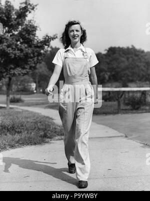1940s HAPPY SMILING WOMAN FEMALE WORKER OUTDOOR WALKING TOWARD  WEARING BIB OVERALLS COVERALLS WORK CLOTHES LOOKING AT CAMERA - q43287 CPC001 HARS GROWING YOUNG ADULT PLEASED JOY LIFESTYLE FEMALES WW2 RURAL GROWNUP HOME LIFE COMMUNICATING FULL-LENGTH LADIES PHYSICAL FITNESS PERSONS OVERALLS GROWN-UP FARMING CONFIDENCE AGRICULTURE B&W EYE CONTACT OCCUPATION CHEERFUL STRENGTH TOWARD WORLD WARS INDEPENDENT WORLD WAR WORLD WAR TWO WORLD WAR II SMILES GROW JOYFUL ROSIE THE RIVETER STYLISH WORLD WAR 2 BIB COMMUNICATE YOUNG ADULT WOMAN BLACK AND WHITE CAUCASIAN ETHNICITY COVERALLS OLD FASHIONED Stock Photo