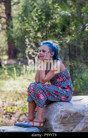 Atrtractive 8 year old girl, in colorful jump suit, hair is dyed bright blue, sitting on rock, in deep thought, side profile. . Model Release #113 Stock Photo