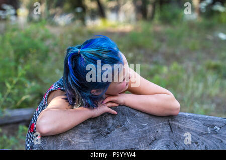 Atrtractive 8 year old girl, in colorful jump suit, hair is dyed bright blue, leaning on bench, side profile. Model Release #113 Stock Photo