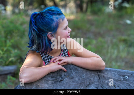 Atrtractive 8 year old girl, in colorful jump suit, hair is dyed bright blue, leaning on bench, side profile. Model Release #113 Stock Photo
