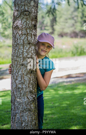 Pretty blonde girl, wearing baseball hat, peeking out from behind a tree. Model Release #113 Stock Photo