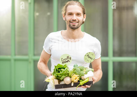 Portrait of a handsome man with boxes full of fresh raw vegetables standing outdoors on the green wall background Stock Photo