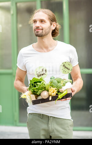 Portrait of a handsome man with boxes full of fresh raw vegetables standing outdoors on the green wall background Stock Photo