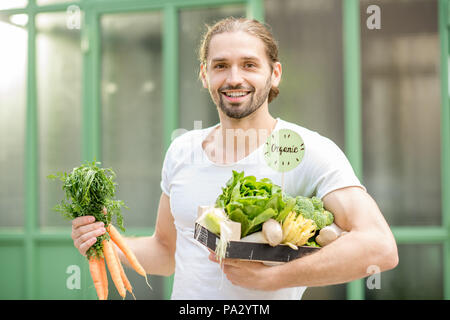 Portrait of a happy vegetarian man holding box full of fresh raw vegetables outdoors on the green background Stock Photo