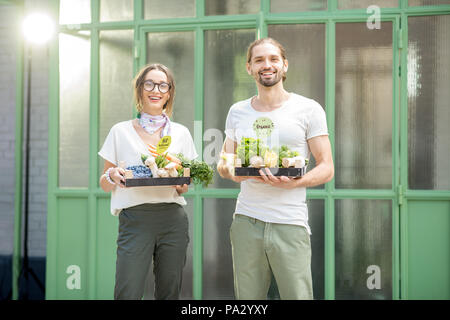 Portrait of a happy vegetarian couple holding boxe full of fresh raw vegetables outdoors on the green background Stock Photo