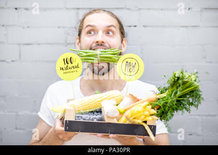 Portrait of a handsome man biting green beans standing with box full of fresh vegetables on the brick wall background Stock Photo