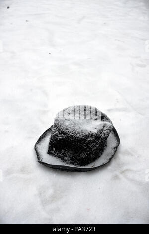an old battered black felt top hat lies in the snow slowly being covered by fresh falling snow on a winter’s day Stock Photo