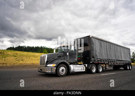Black stylish big rig American popular professional semi truck for local haul delivery transporting cargo inside covered black semi trailer with spoil Stock Photo
