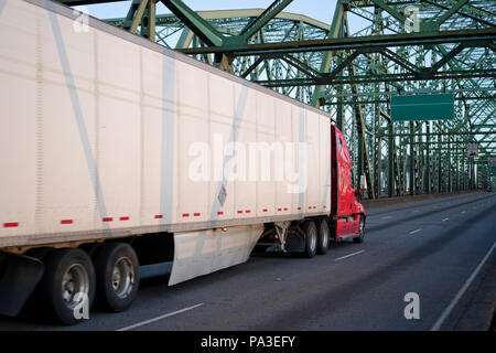 Red big rig long haul semi truck driving alone interstate highway transporting packed commercial cargo in dry van semi trailer moving on arched metal  Stock Photo