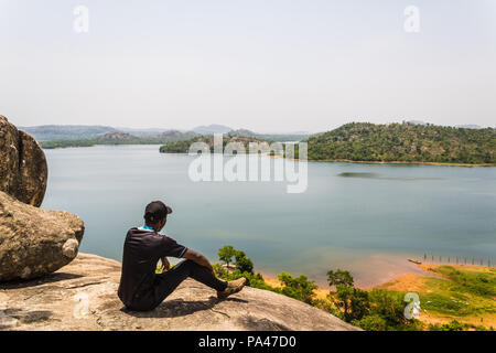 Sitting on an outcropped rock on the top of a mountain looking out towards the lake and island. Stock Photo