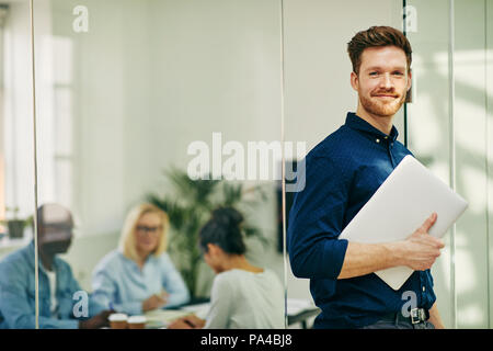 Smiling young businessman standing at the door of an office boardroom holding a laptop with colleagues at work in the background Stock Photo