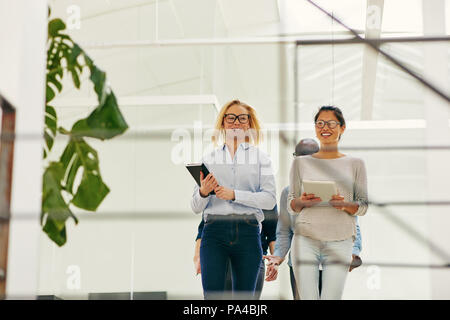Two smiling young businesswomen talking together while walking with male colleagues to a meeting in a bright modern office Stock Photo