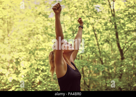 Fit young woman in sportwear smiling while standing on a path with her arms raised in victory after a run through the woods Stock Photo