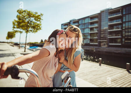 Two young girlfriends laughing together while walking arm in arm with a bicycle through the city in summer Stock Photo