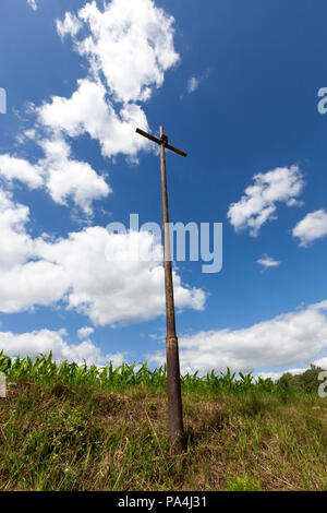 a tall metallic religious old cross set on a hill in a field, against a blue cloudy sky and green grass Stock Photo