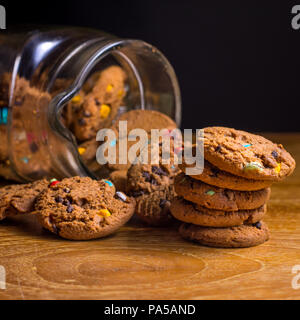 Choc chip smartie cookies falling out of jar on wooden table top. Stock Photo