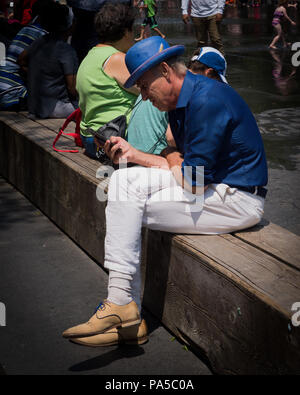 Man wearing blue derby hat, electric blue shirt, white pants reading cell phone while staying cool in hot summer weather. Stock Photo